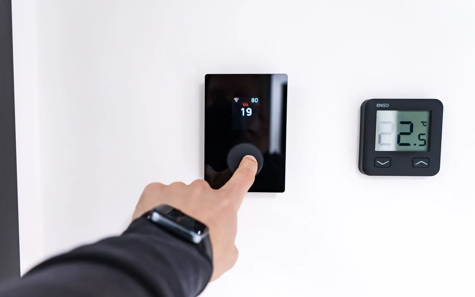 A hand pressing the dial on a digital heating control mounted on a wall
