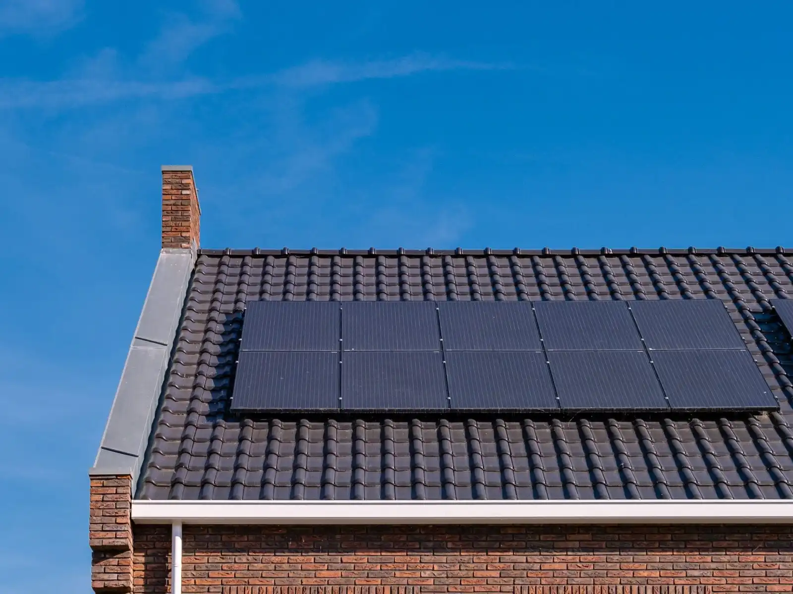 Black solar panels on the dark tiled roof of a brick house, with a chimney and a clear blue sky