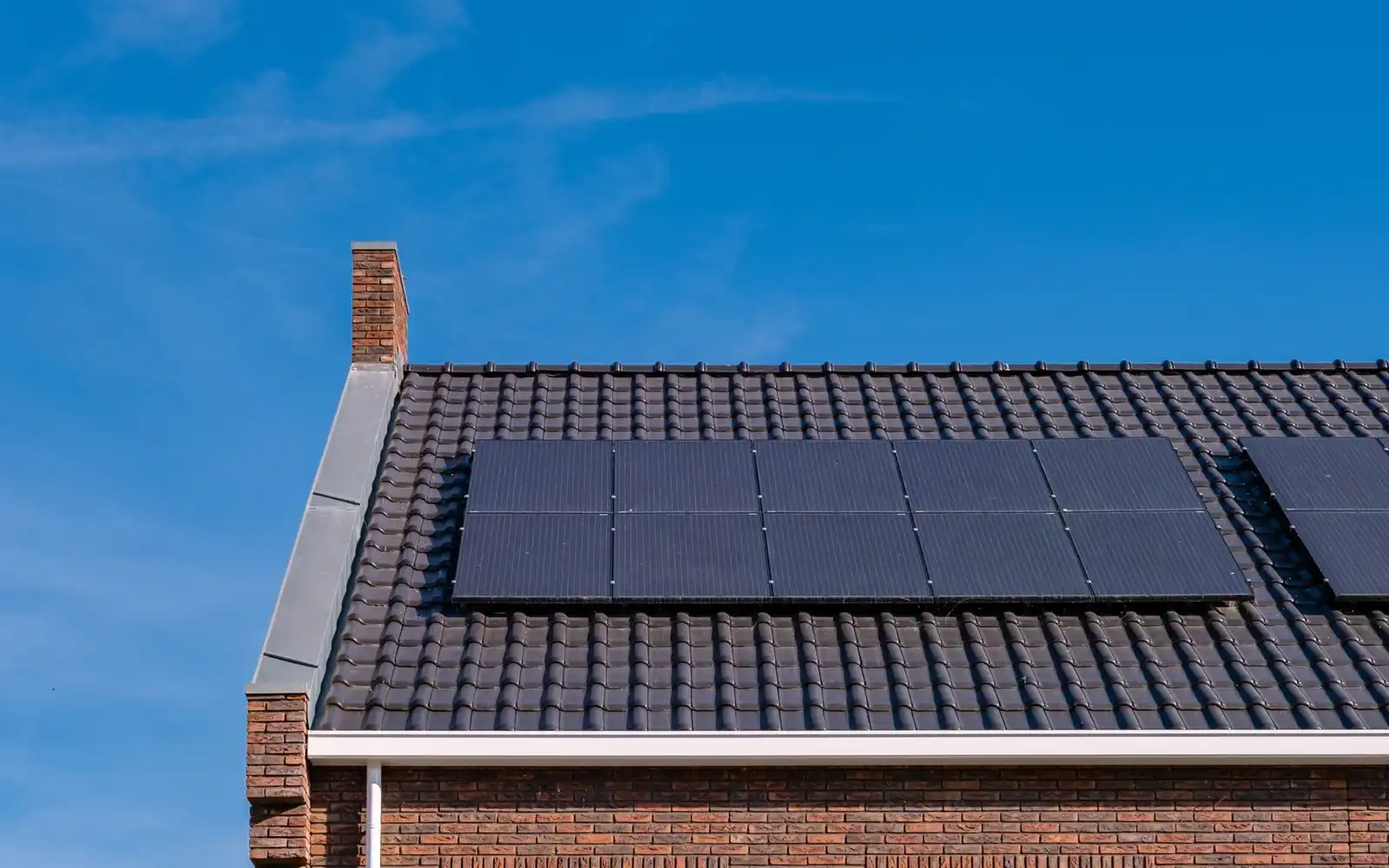 Black solar panels fitted flush to the dark tiled roof of a brick house against a clear sky